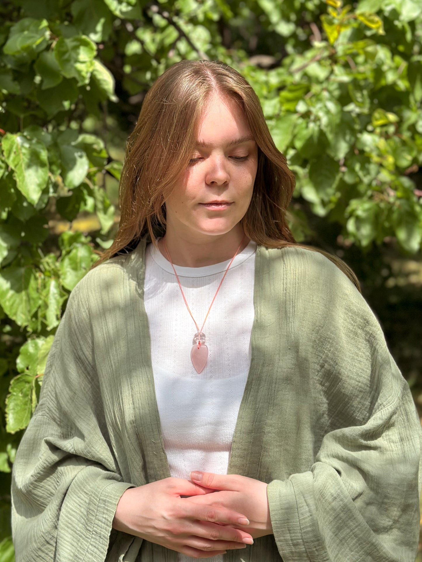 Woman wearing a green cardigan and white shirt with a pink pendant, standing in front of green foliage.