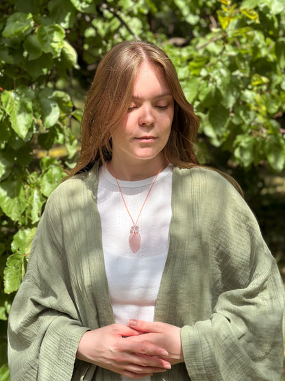 Woman wearing a green cardigan and white shirt with a pink pendant, standing in front of green foliage.