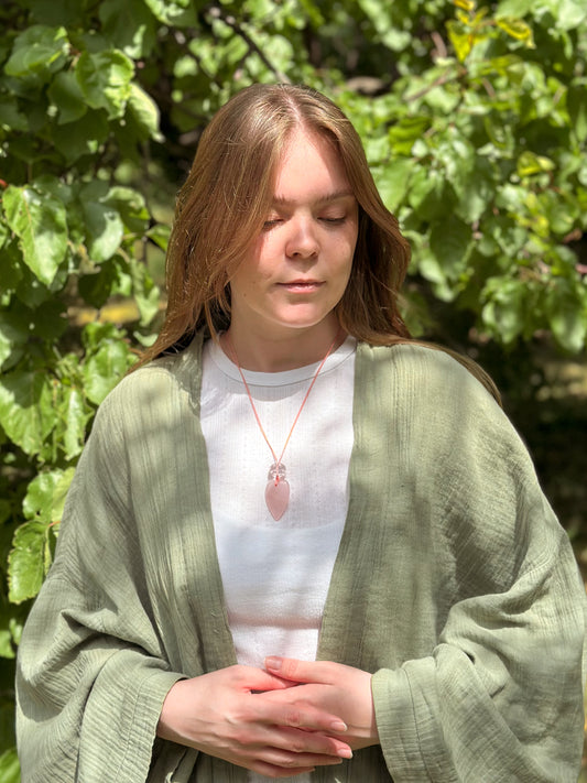 Woman wearing a green cardigan and white shirt with a pink pendant, standing in front of green foliage.