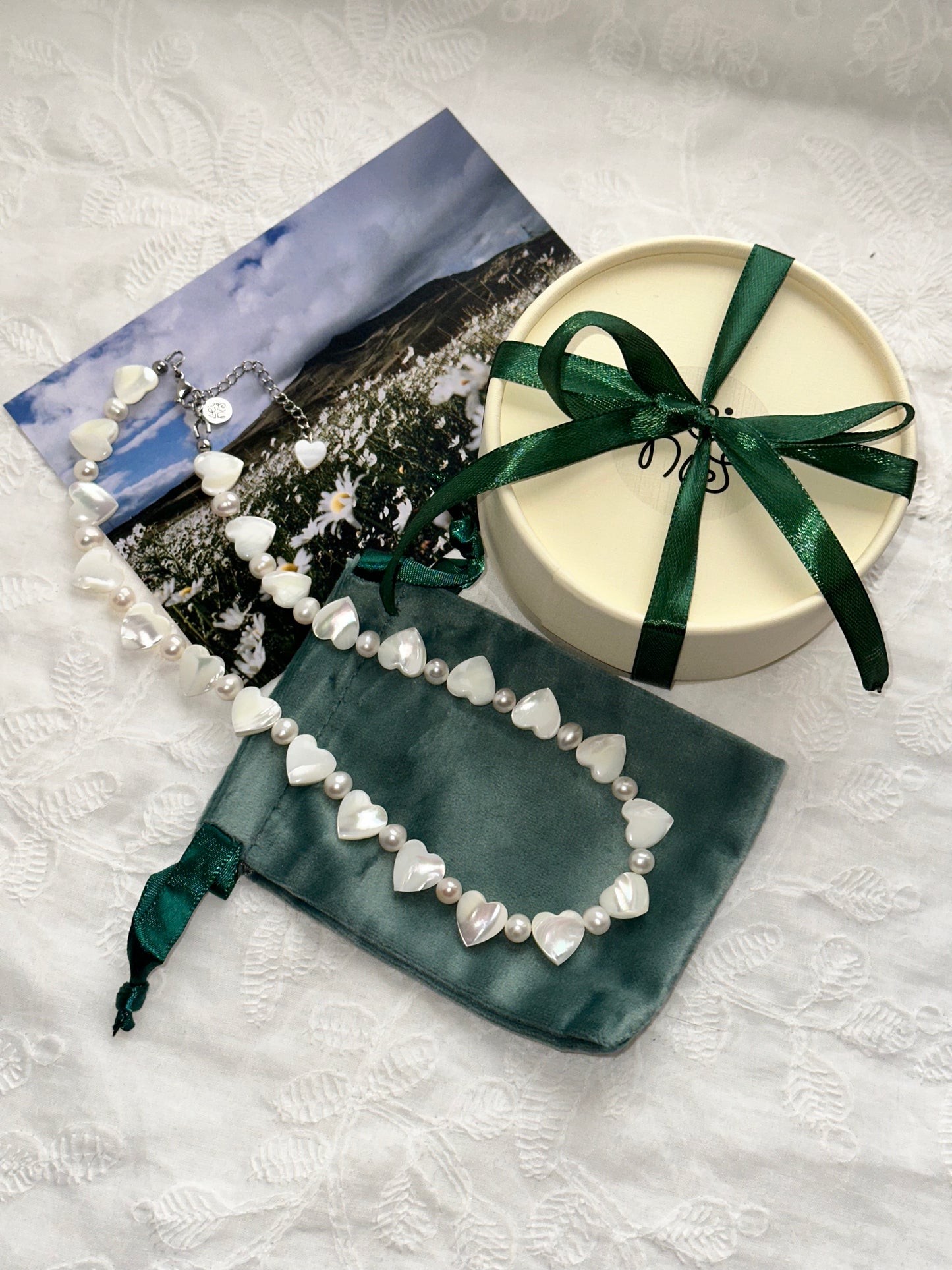 Pearl necklace on a green velvet bag with a scenic card and round box with a green ribbon on a textured white surface.