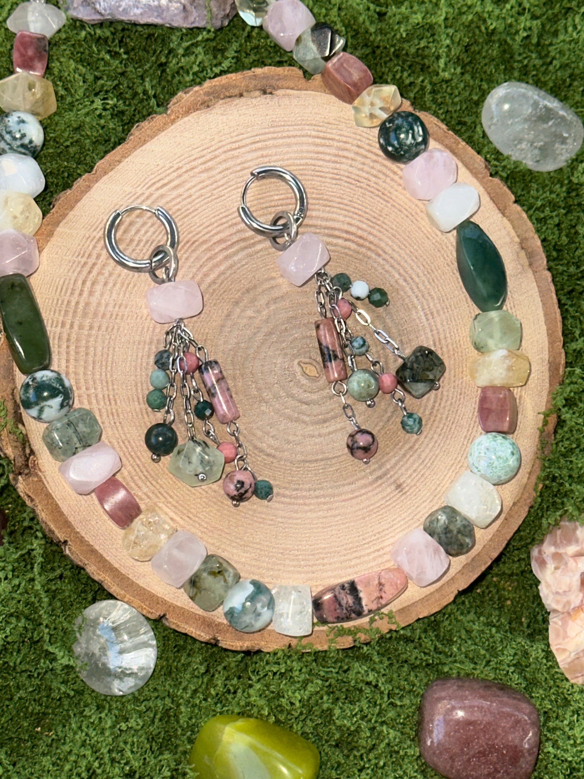 Pair of earrings with gemstones on a wooden slice surrounded by more gemstones on a grassy background