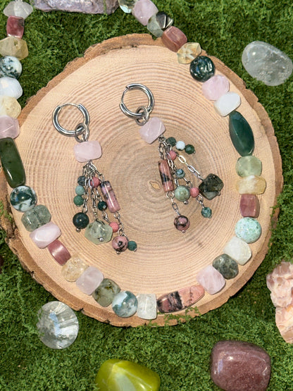 Pair of earrings with gemstones on a wooden slice surrounded by more gemstones on a grassy background