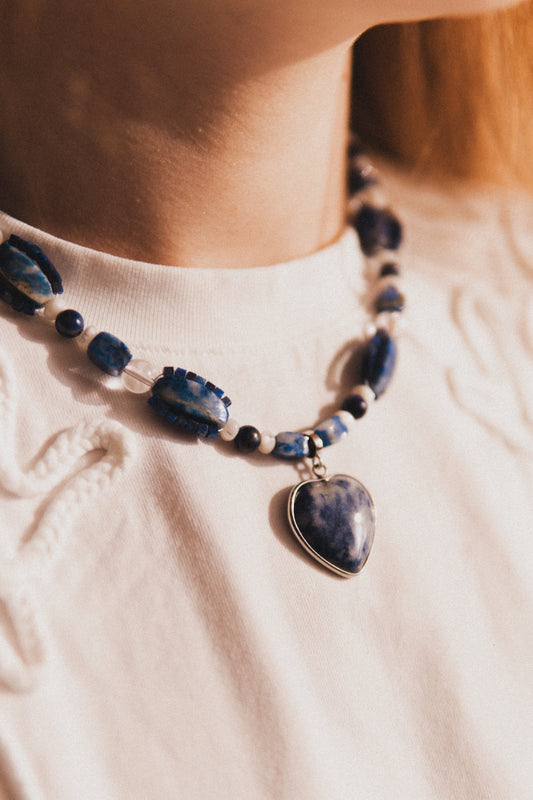 Close-up of a blue beaded necklace with a heart pendant on a white shirt.