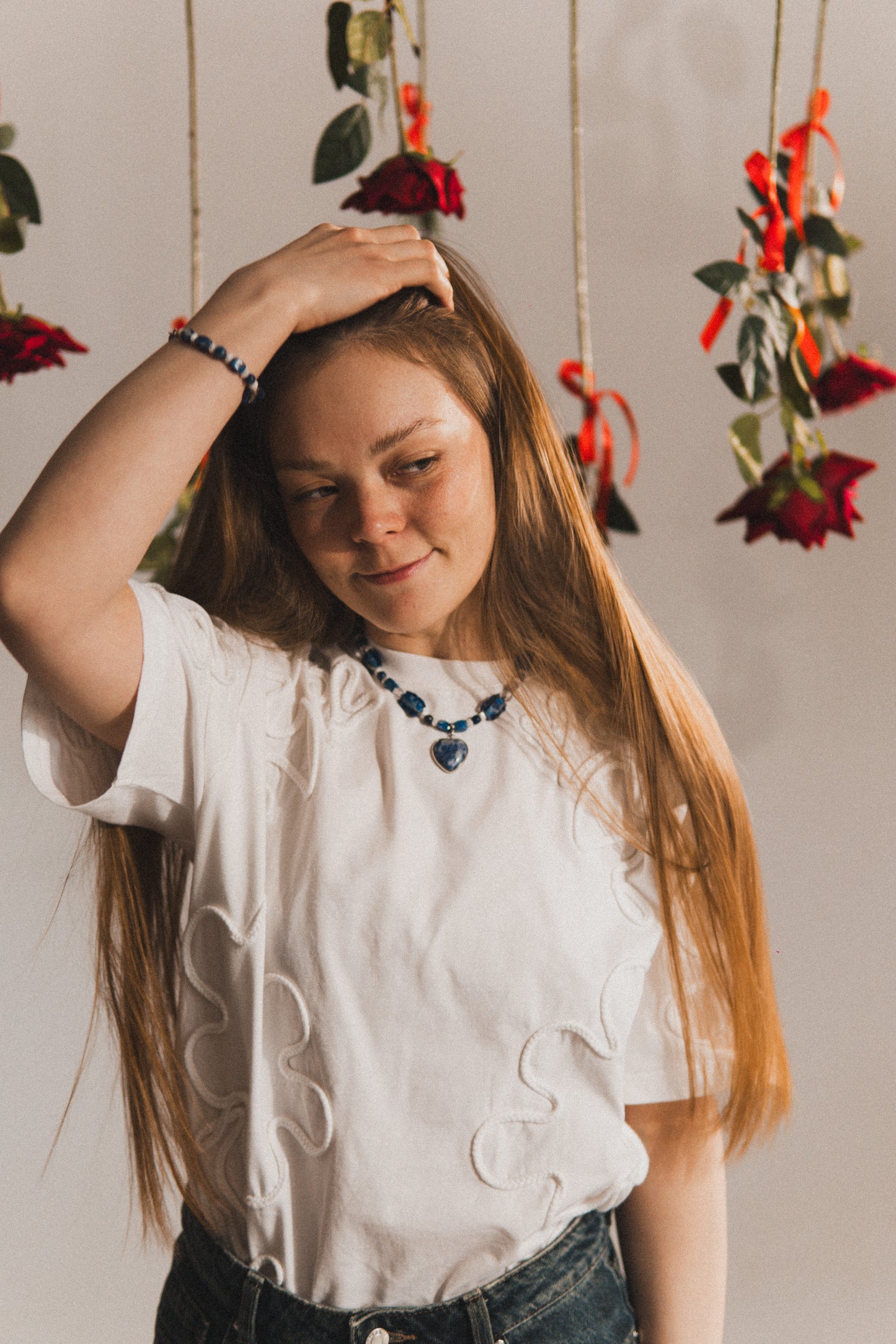 Woman with long hair wearing a white blouse and dark necklace, standing against a neutral background with hanging floral decorations.