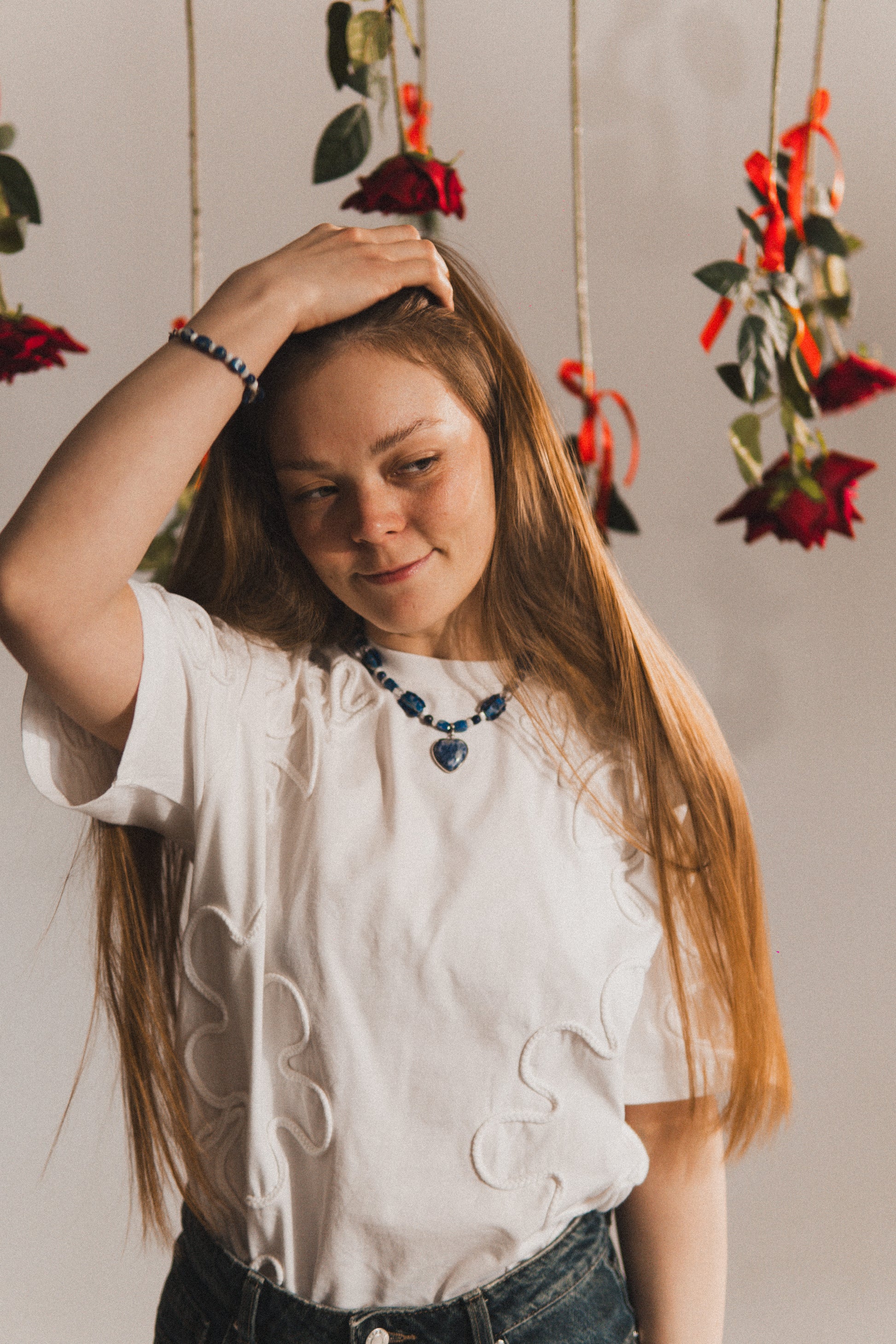 Woman with long hair wearing a white blouse and dark necklace, standing against a neutral background with hanging floral decorations.
