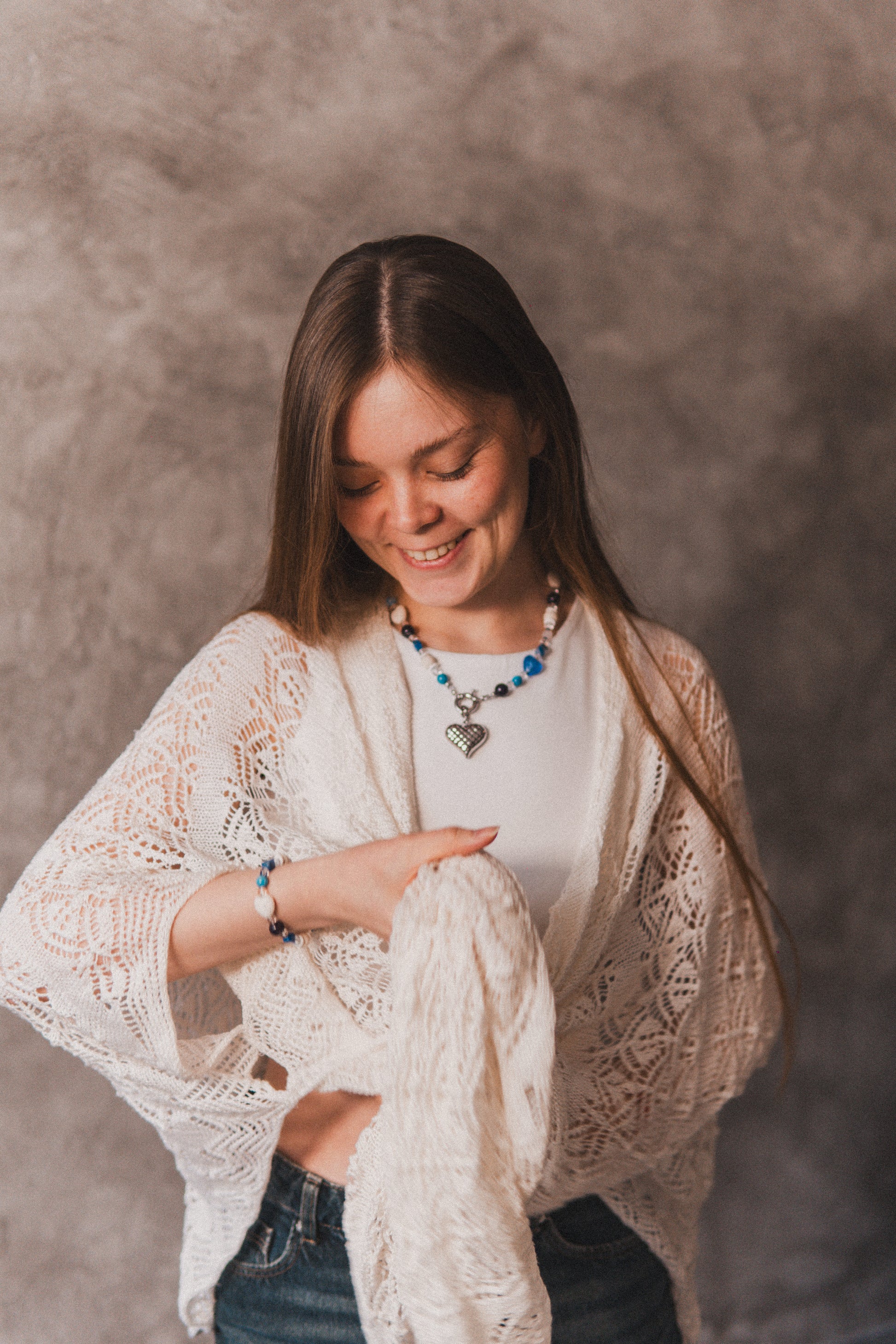 Woman wearing a white lace cardigan over a white shirt with a necklace, sitting against a plain background.