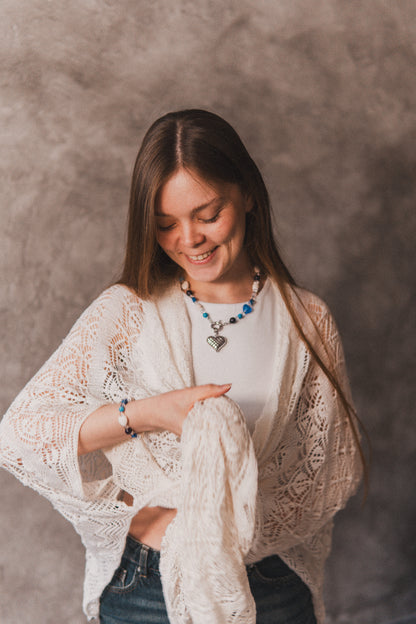 Woman wearing a white lace cardigan over a white shirt with a necklace, sitting against a plain background.