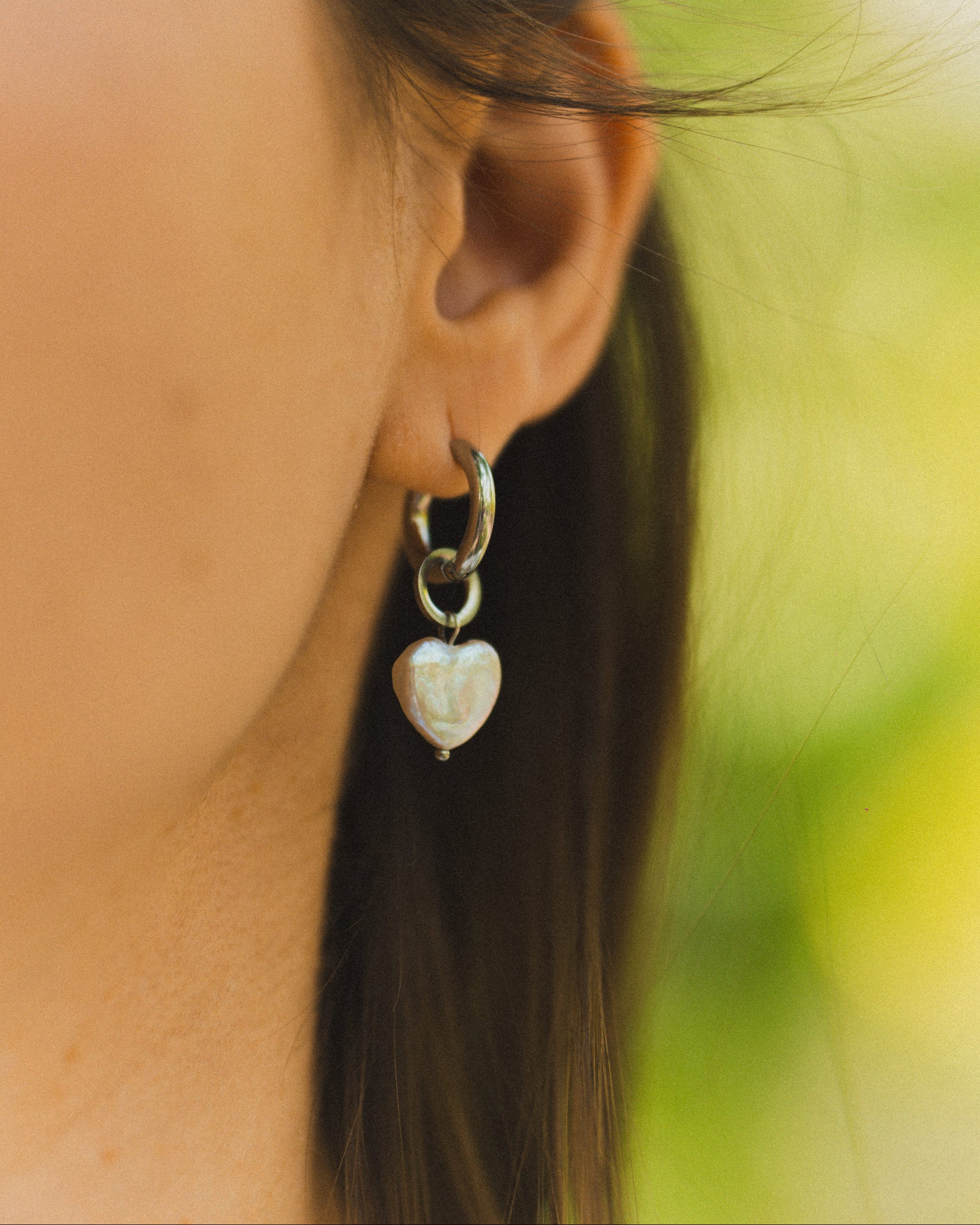 Close-up of a person wearing a silver earring with a heart-shaped pendant against a blurred natural background.
