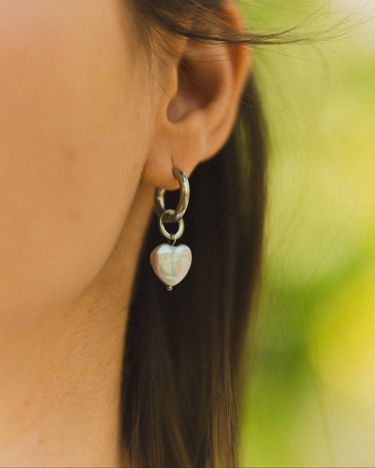 Close-up of a person wearing a silver earring with a heart-shaped pendant against a blurred natural background.